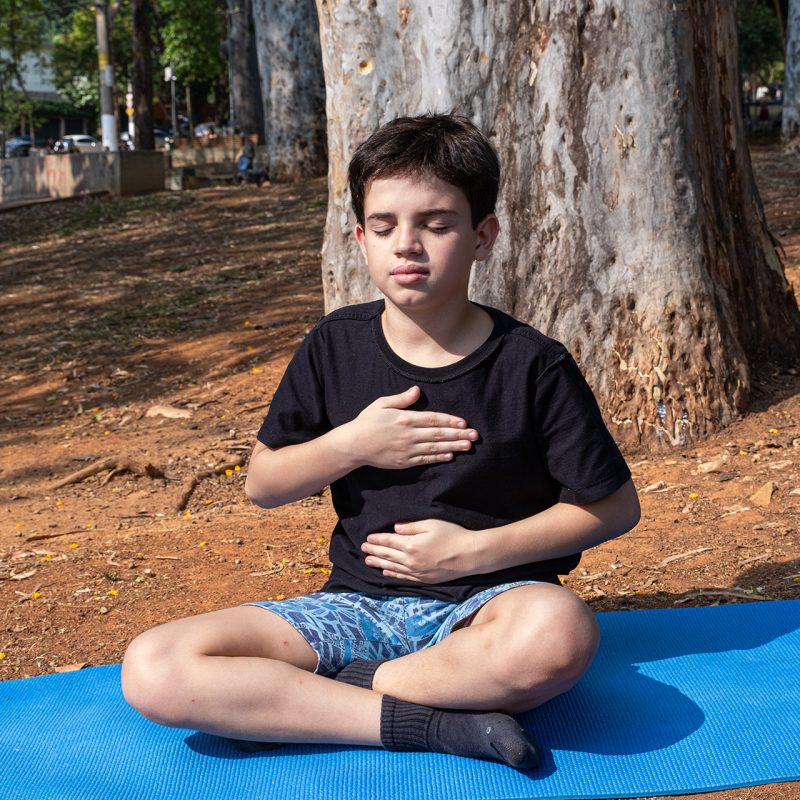 Brazilian child sitting on yoga mat with hands on chest and abdomen in outdoor class_4.
